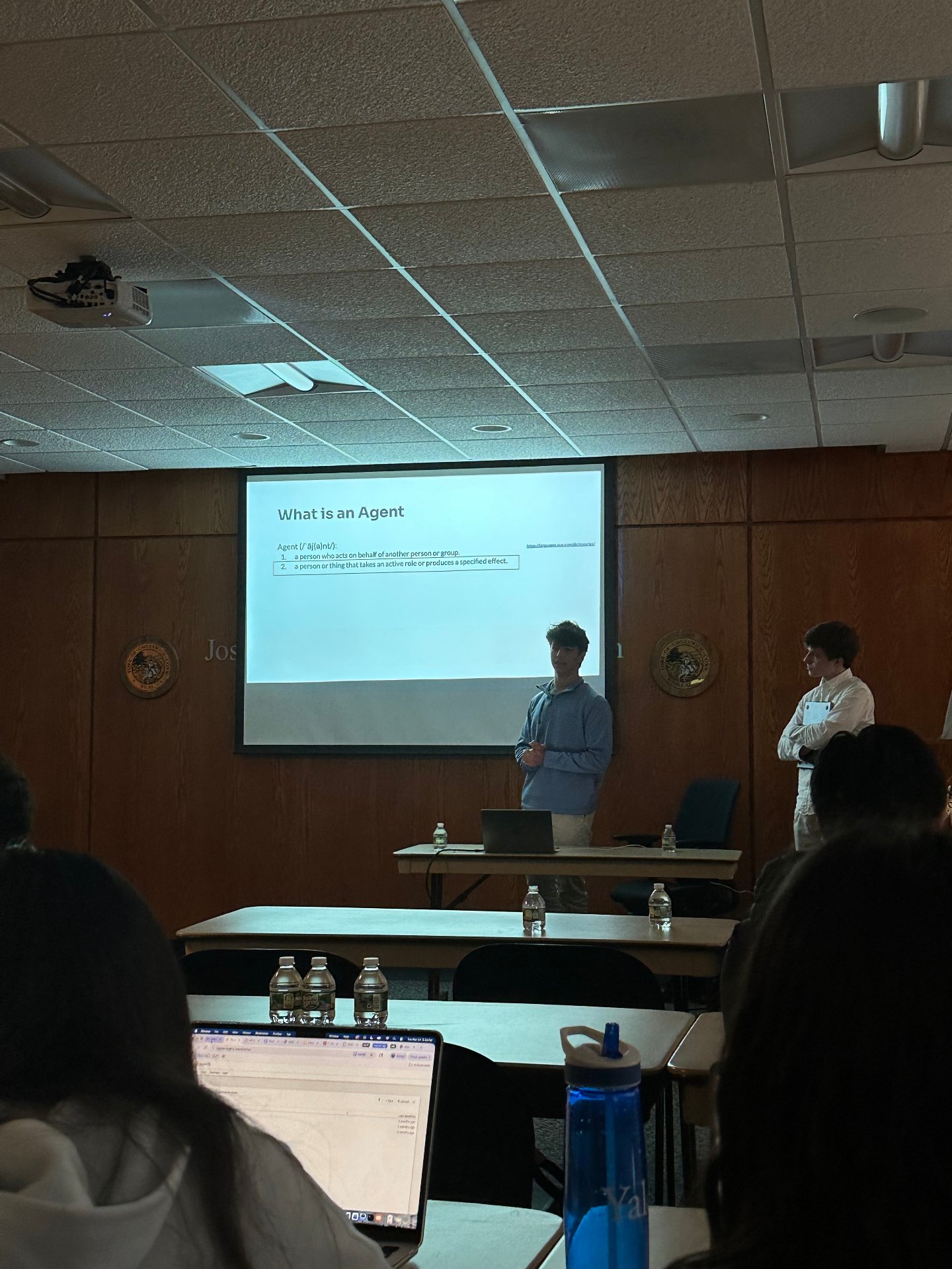 A slide reading What is an Agent projected during the AI in quantitative finance session, with students taking notes on laptops