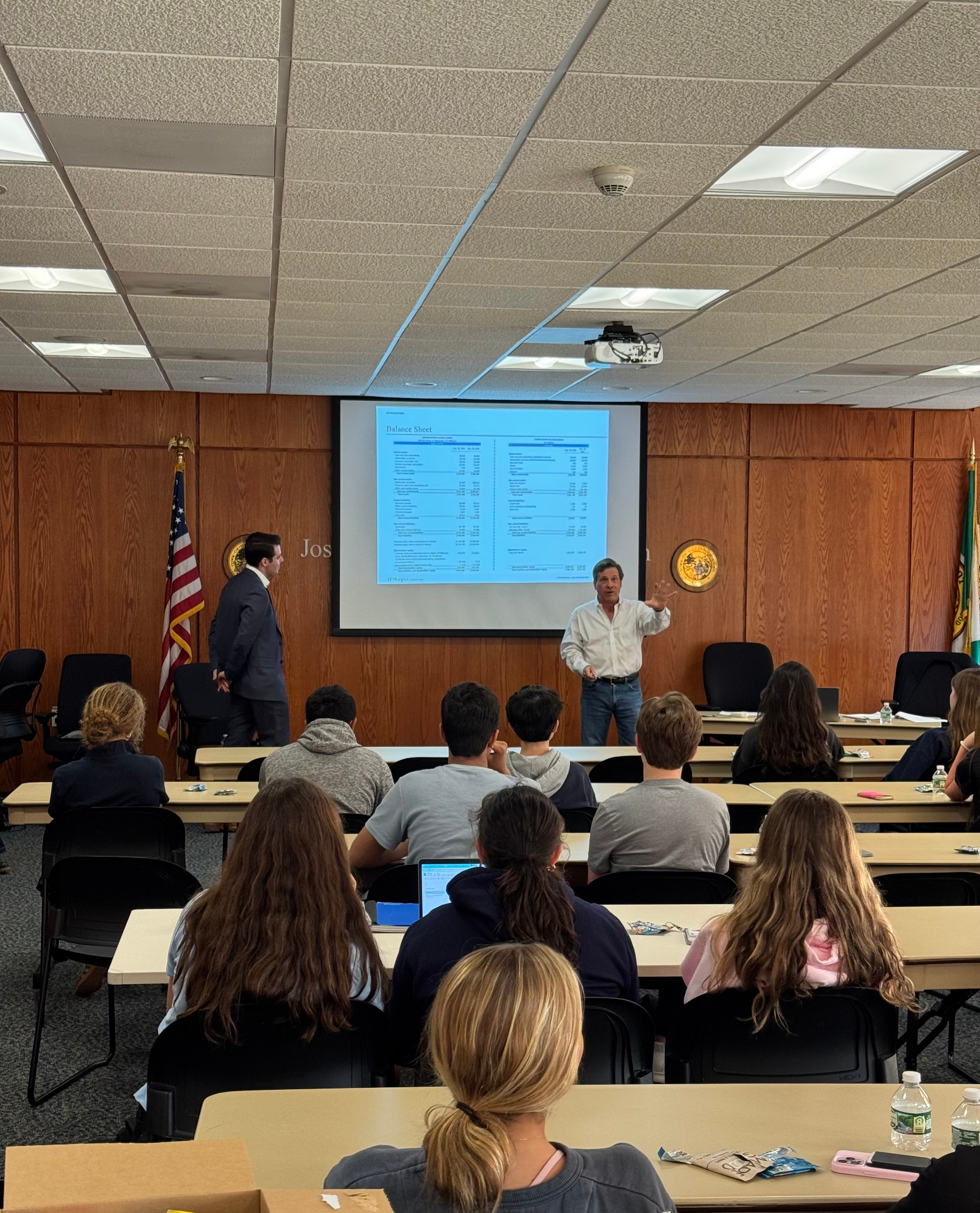 A financial professional in a suit presenting a table of tax figures to seated Greenwich high school students