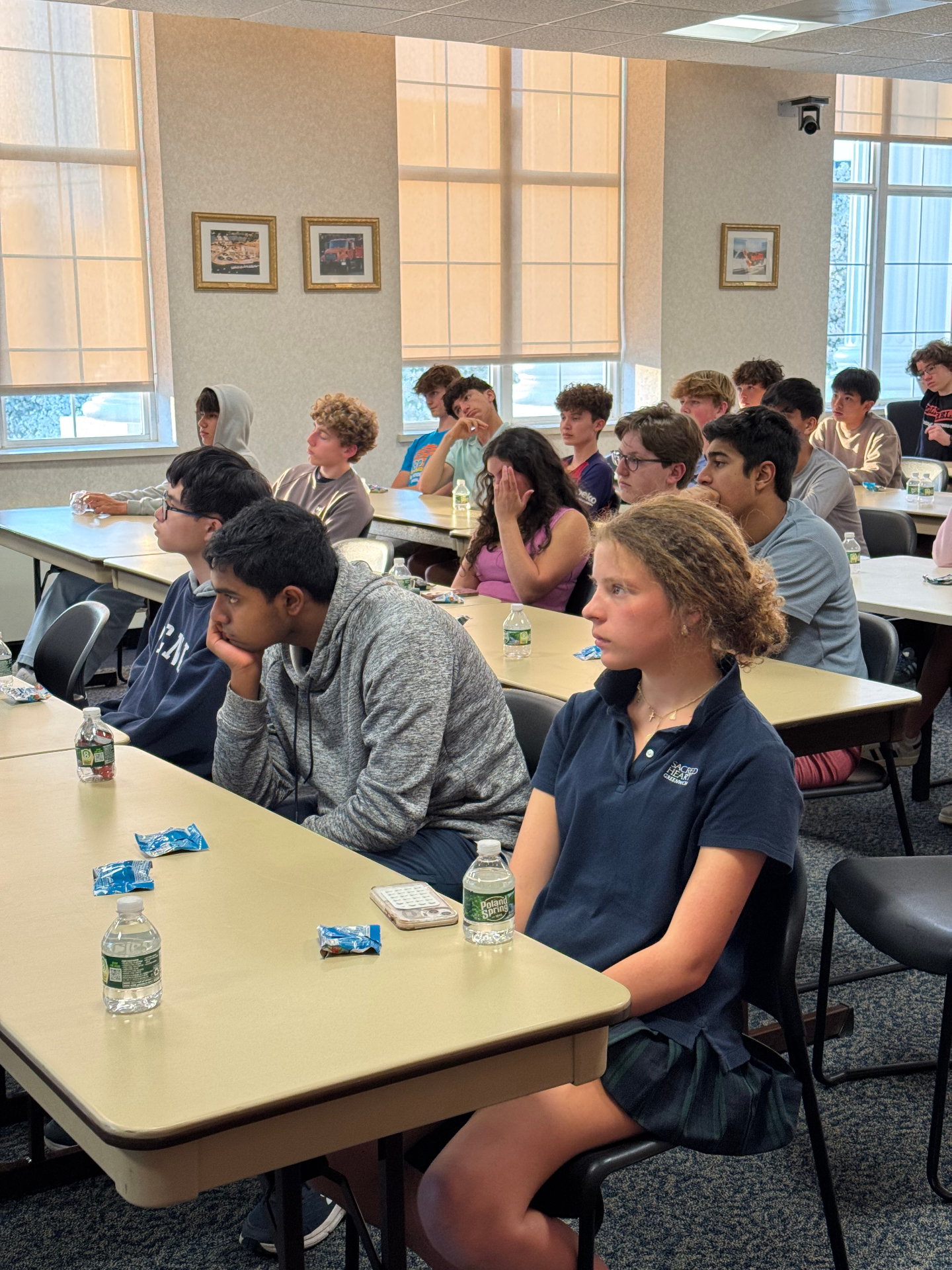 Students in school uniforms listening attentively along a long table near the windows of the meeting room